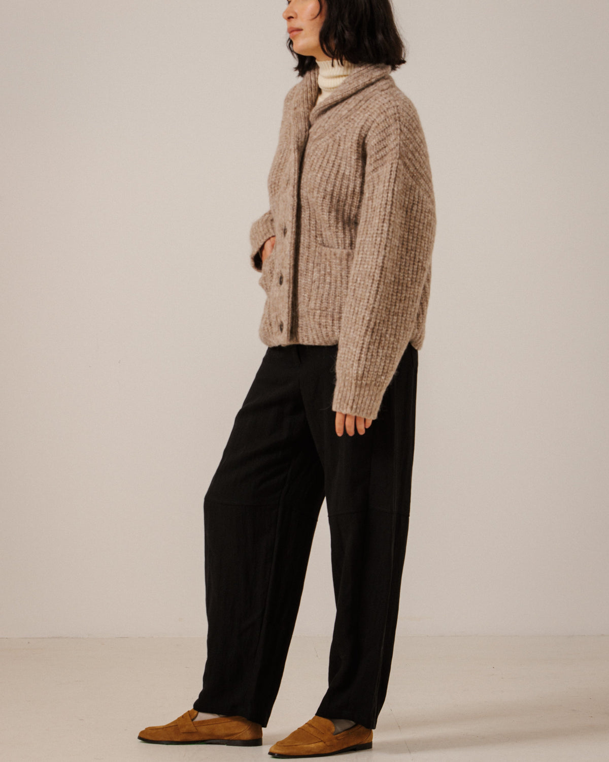 A woman stands in profile wearing a chunky knit beige cardigan over a white turtleneck, black wide-leg trousers, and tan slip-on shoes against a plain light gray studio background.