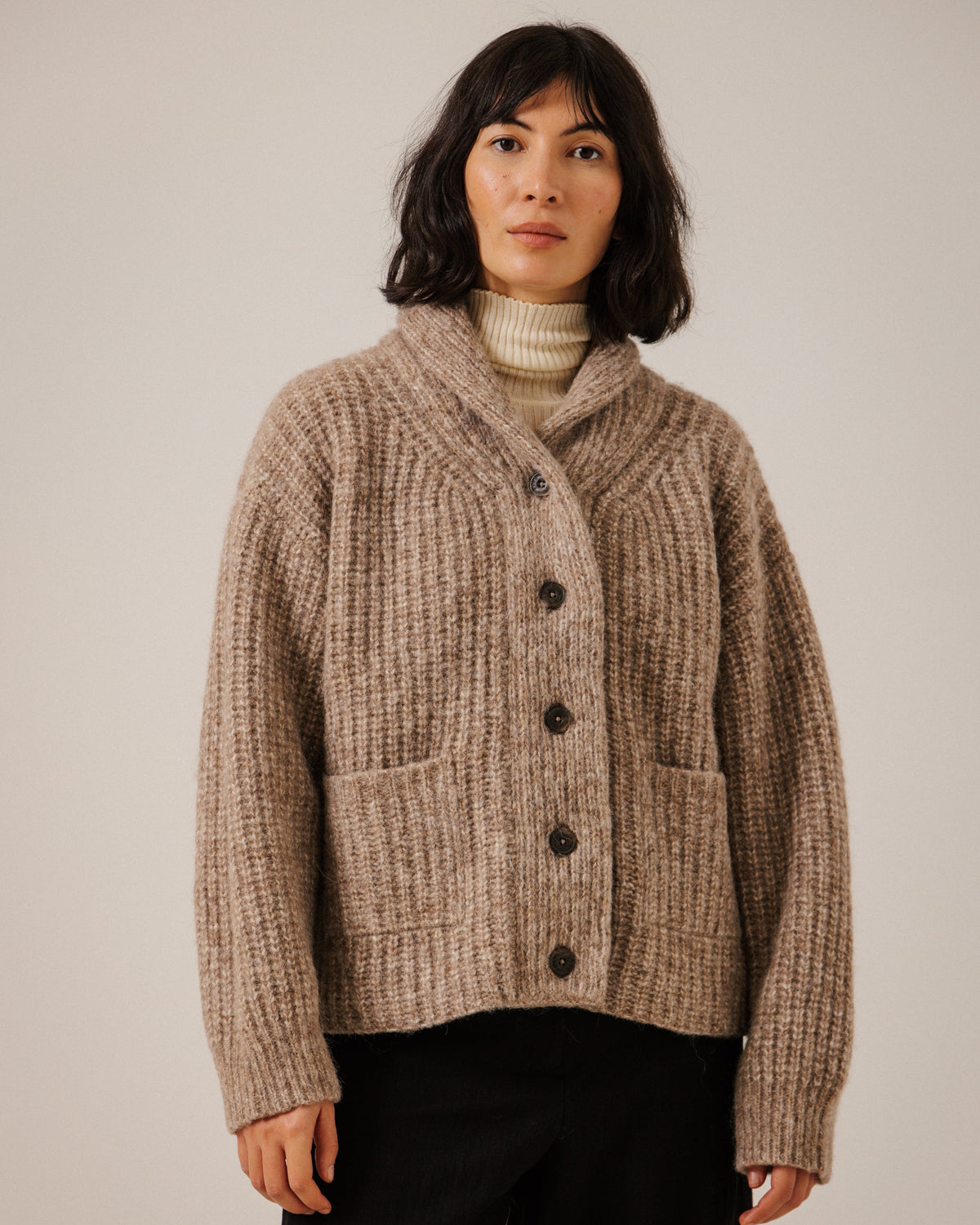 A woman wearing a chunky ribbed knit cardigan in a warm taupe-brown color, styled over a cream turtleneck and black trousers, standing against a plain light gray studio background.