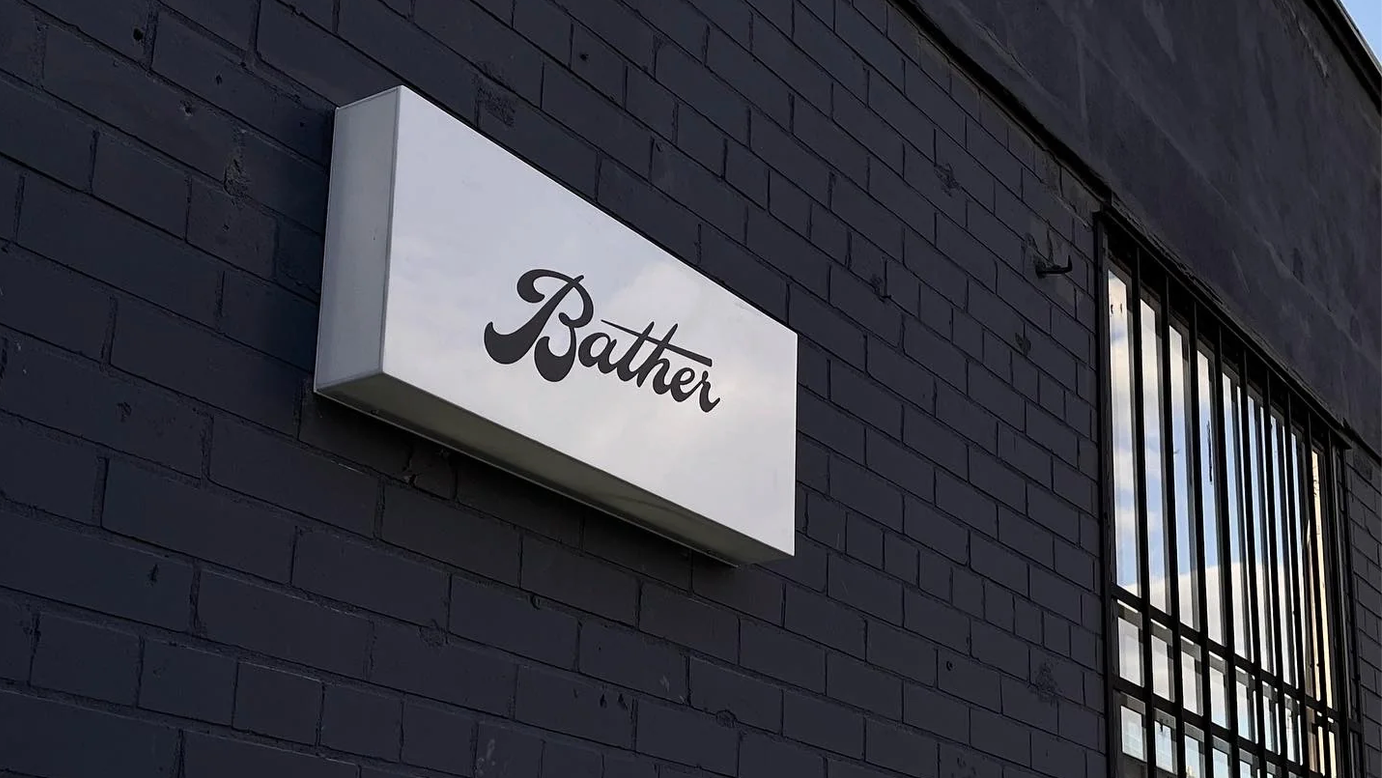 A rectangular white wall sign with the word 'Bather' in black script, mounted on a dark charcoal brick wall next to a barred window.