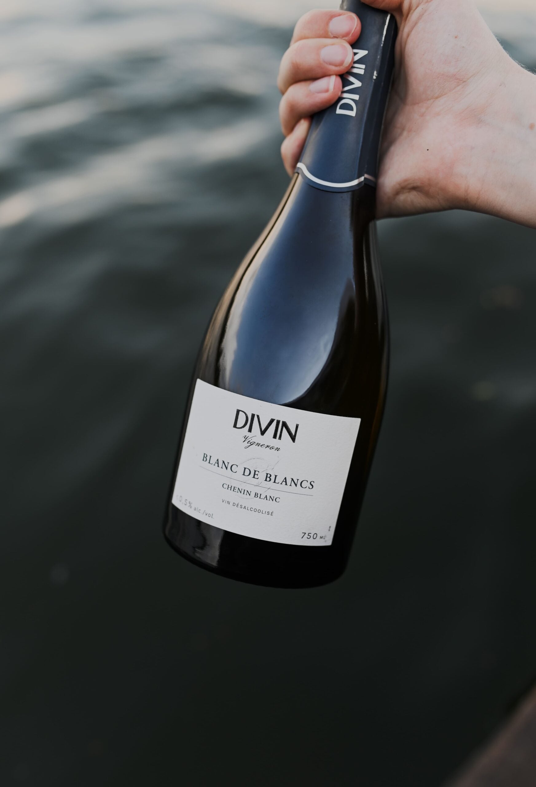 Hand holding a bottle of Divin Blanc de Blancs wine with water in the background