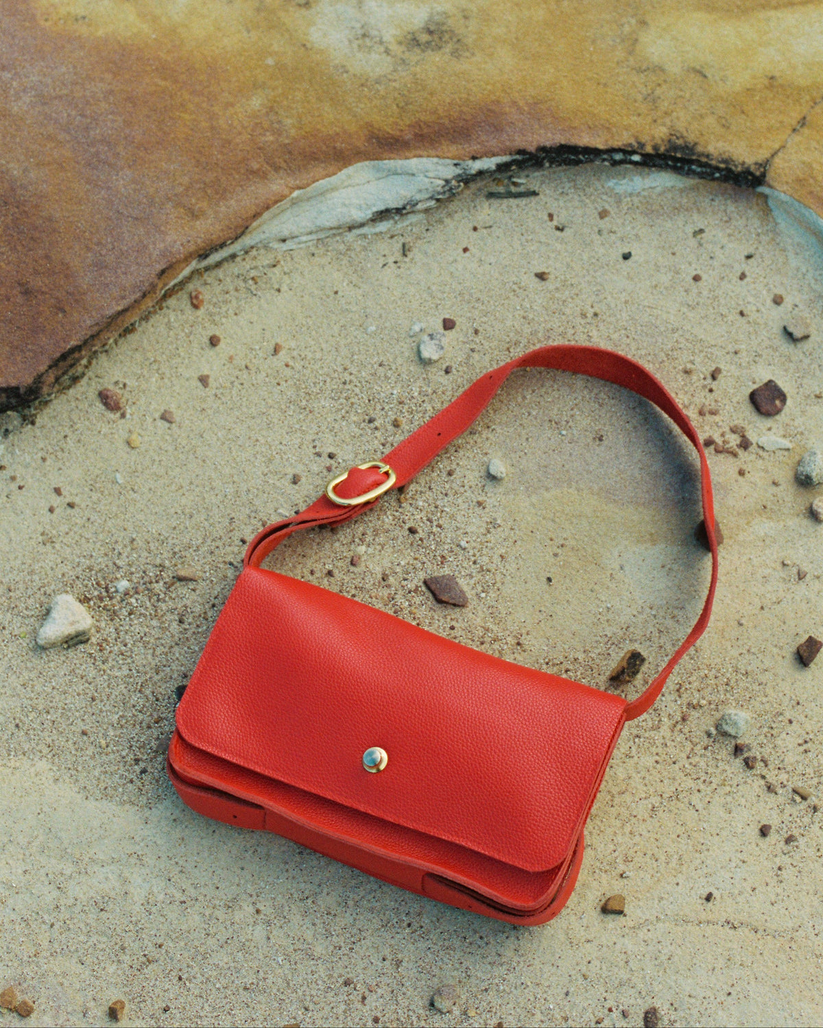 A bright red leather shoulder bag with a flap front and gold-tone hardware, placed on a sandy surface with a large textured rock behind it.