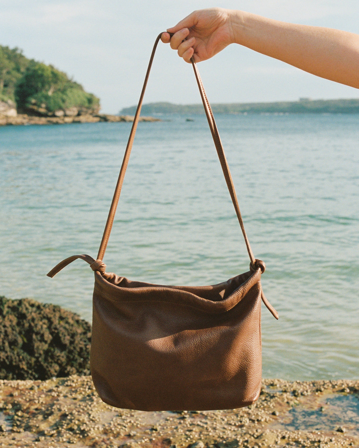 a brown leather hand bag held up in front of beach and ocean