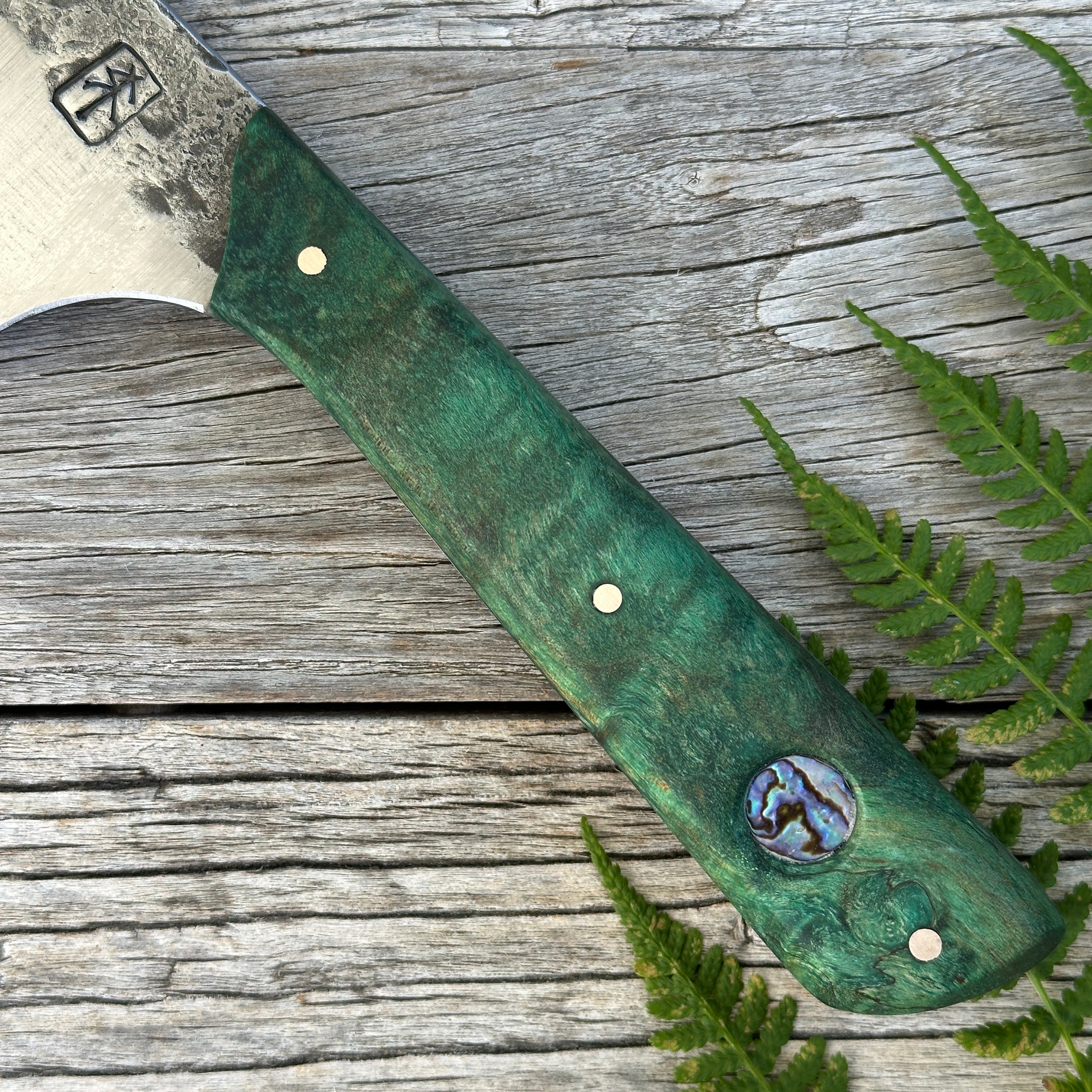 Green-handled Leechtown Blacksmith Co. for Confluence Nimmo Bay knife with a decorative head on a wooden surface with ferns.