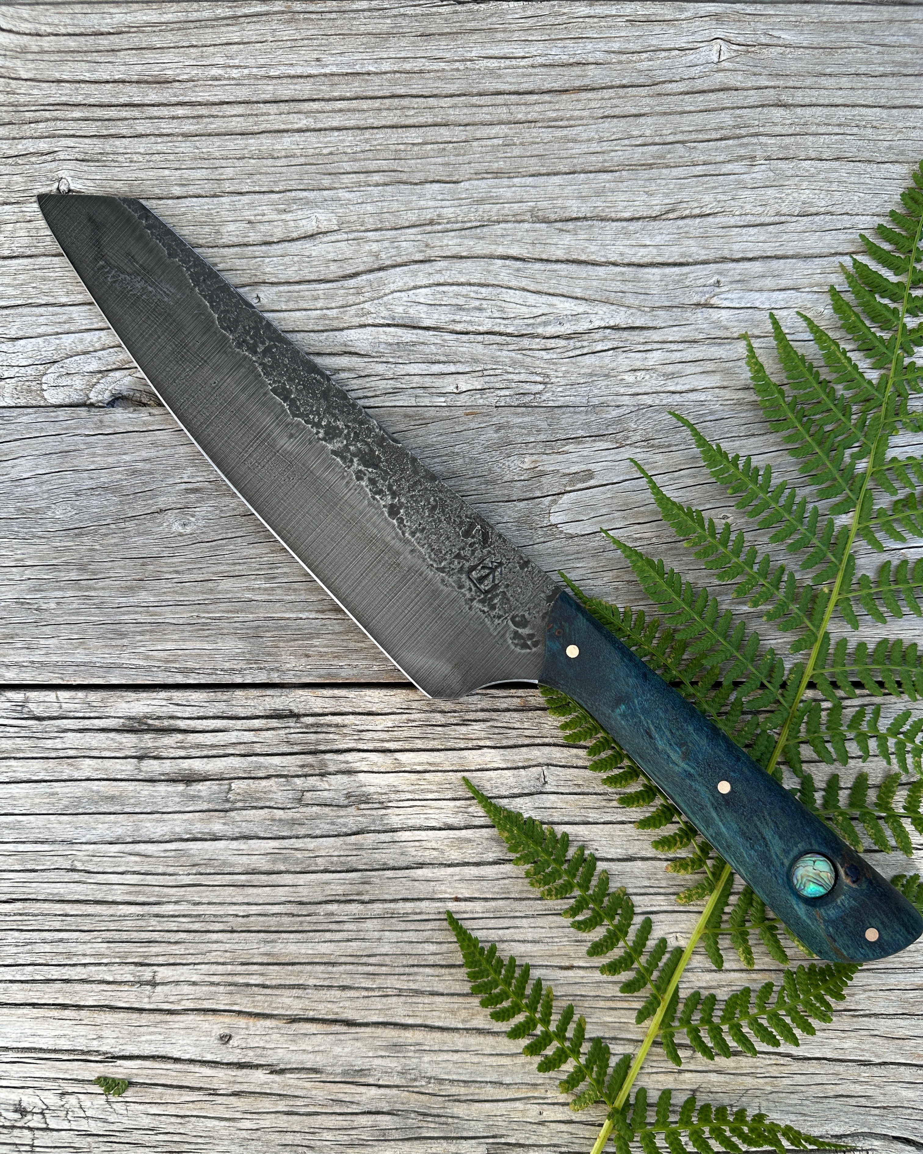 Leechtown Blacksmith Co. for Confluence Nimmo Bay knife with a textured blade and blue handle on a wooden surface with fern leaves.