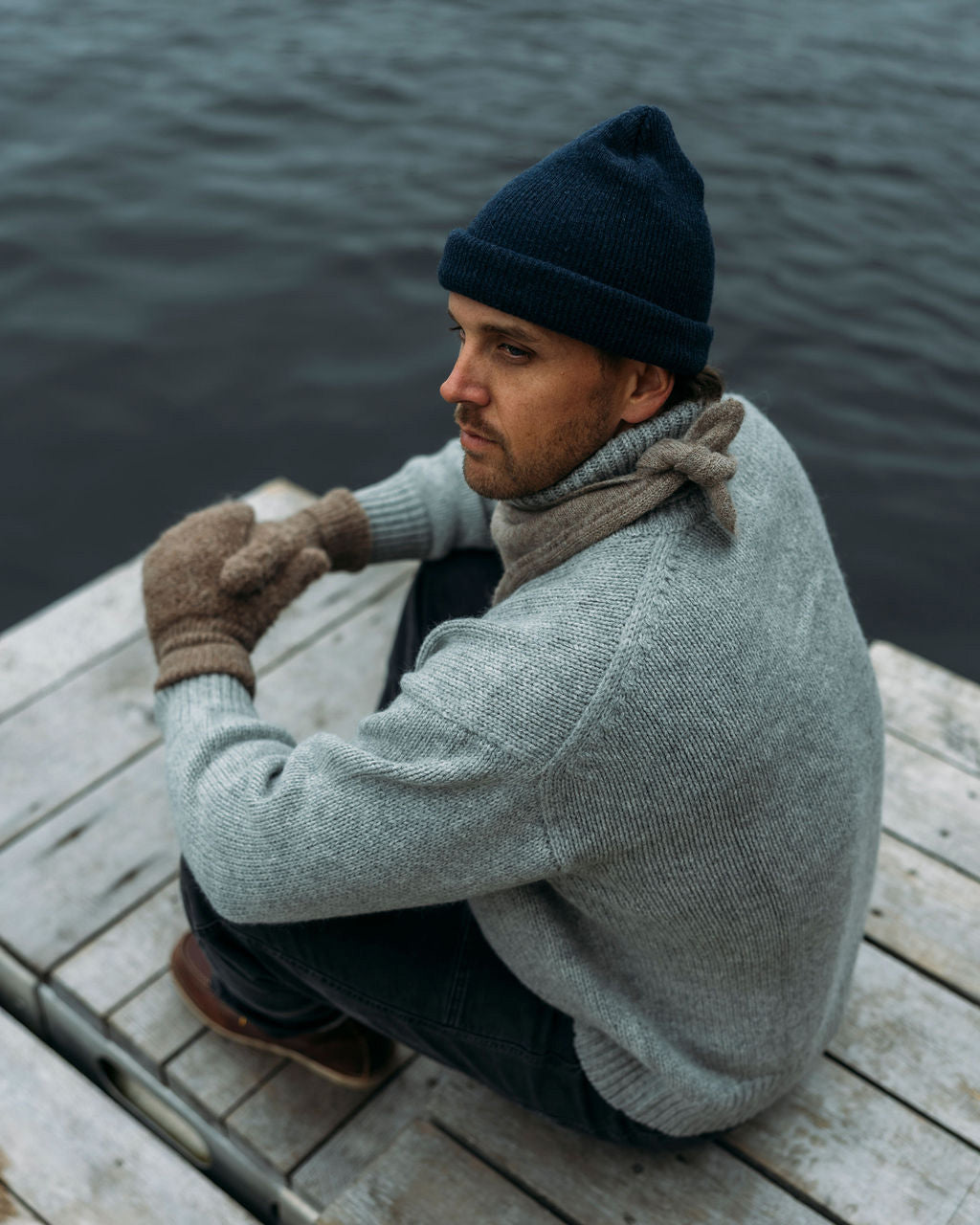 Man sitting on a dock wearing a gray sweater, blue beanie, and Bare Knitwear mittens. 