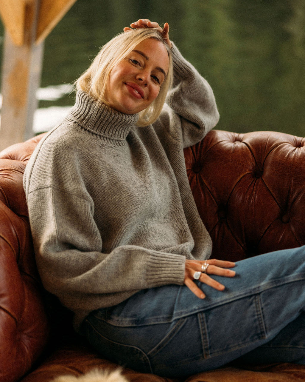 Woman wearing a gray Bare Knitwear turtleneck sweater and blue jeans sitting on a brown leather couch.