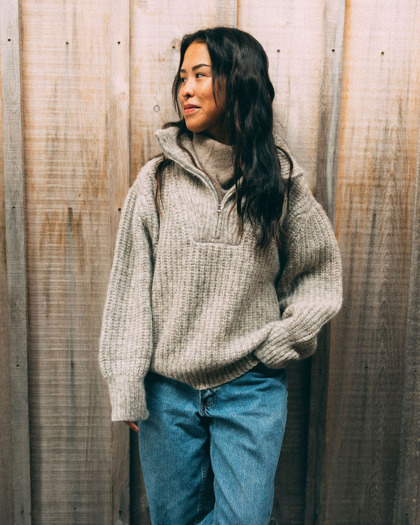 Woman wearing a beige Bare Knitwear 1/4 zip sweater and white skirt standing by a body of water with trees in the background.