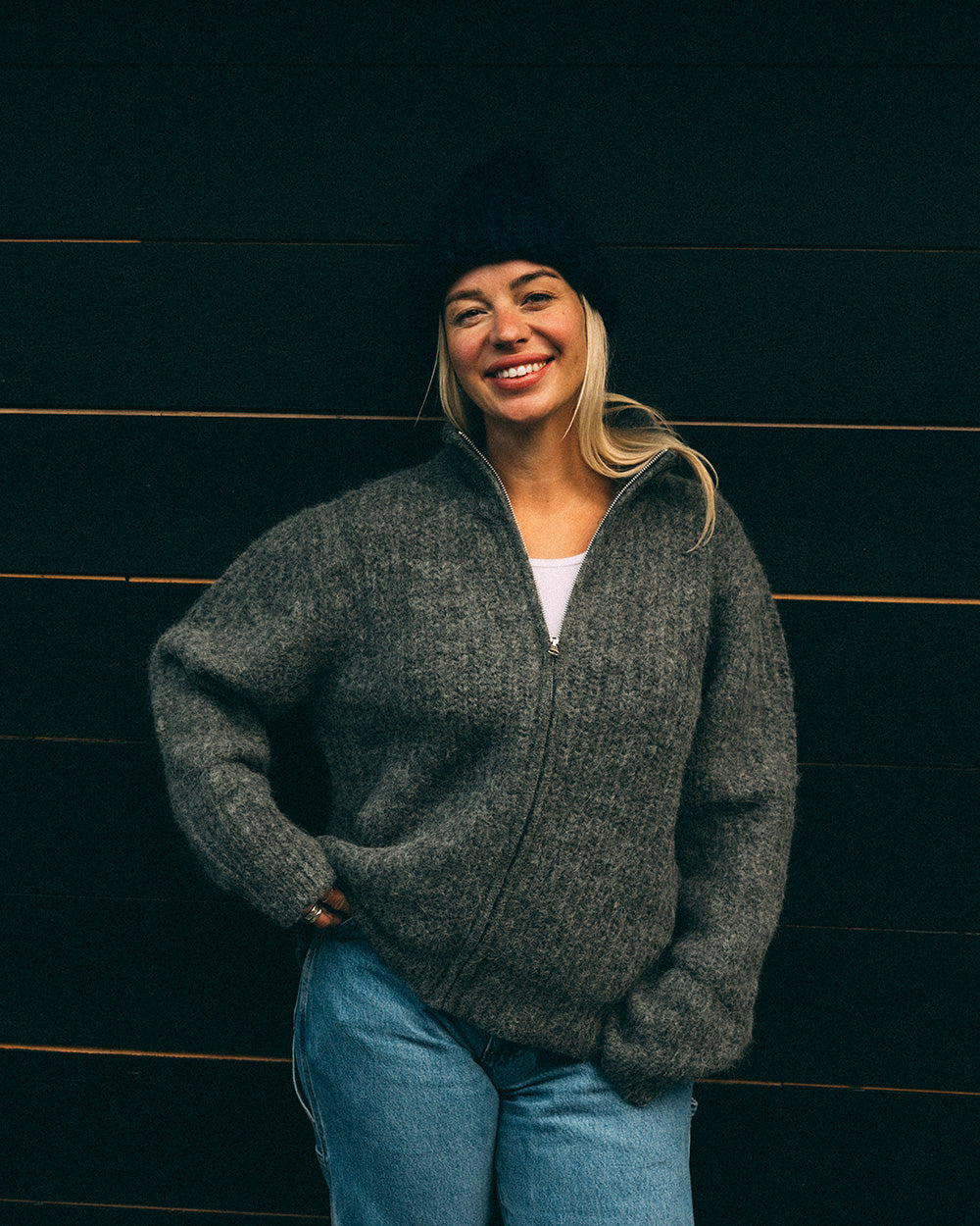 Woman wearing a grey Bare Knitwear full zip sweater and blue jeans against a dark wooden background