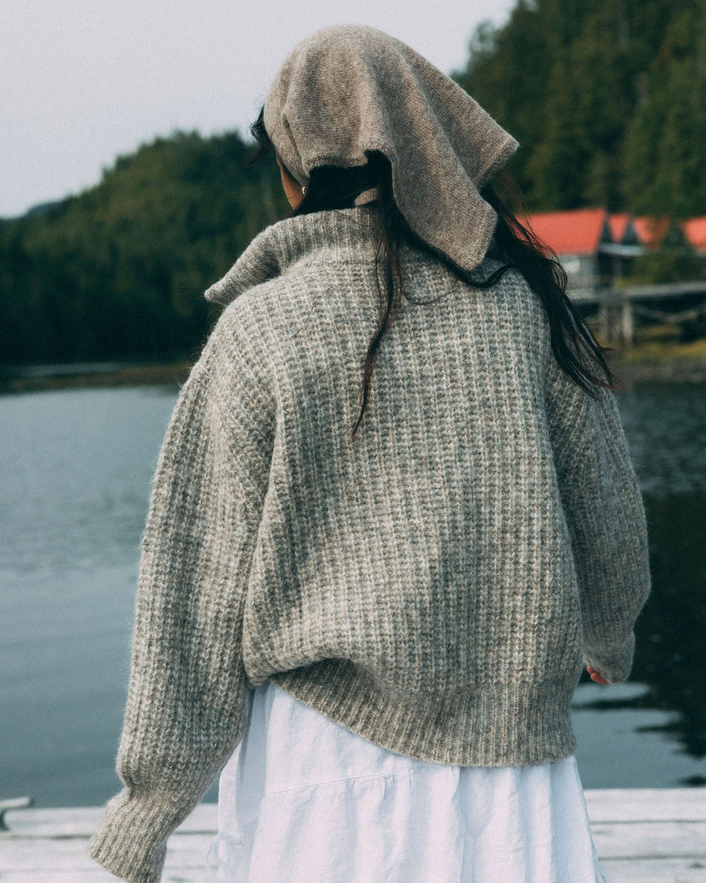 Woman wearing a beige Bare Knitwear 1/4 zip sweater and white skirt standing by a body of water with trees in the background.