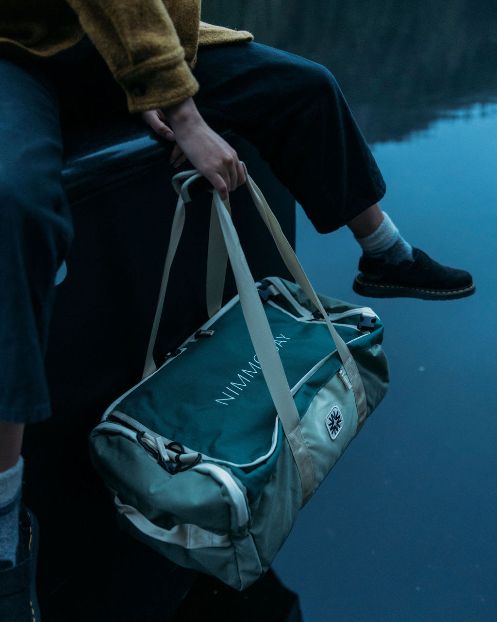 Person holding a green duffel bag with 'Nimmo Bay' branding by a body of water.