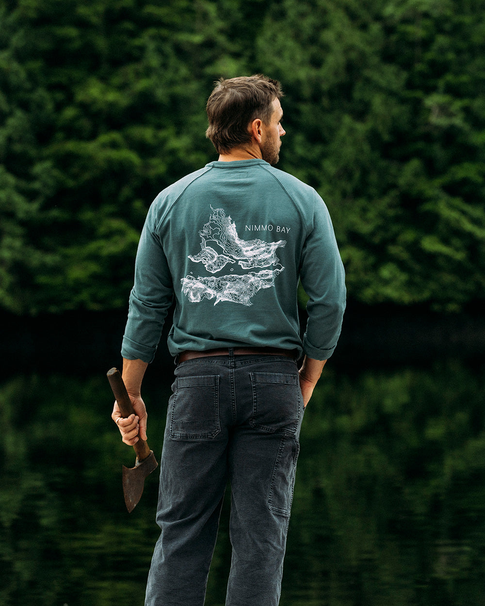 Man wearing a green anian henley with a nimmo bay topical map pn the back, standing in a forest.