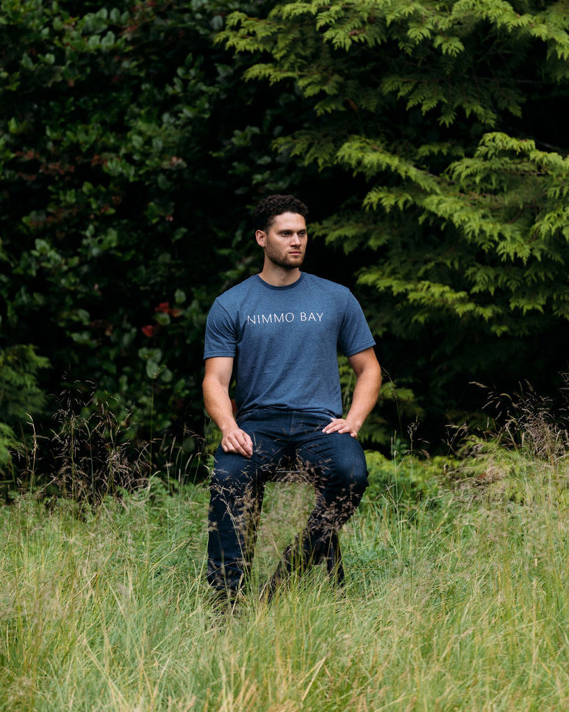 Man wearing a blue 'Nimmo Bay' t-shirt sitting in a grassy area with trees in the background